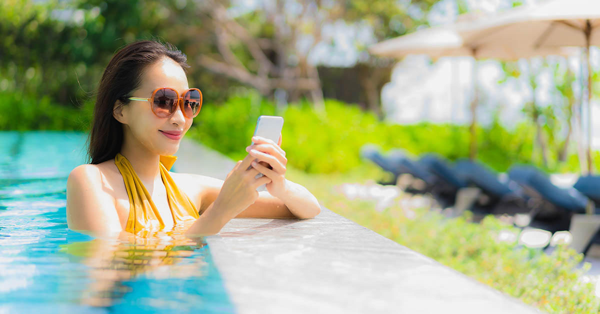 Mulher relaxando na piscina em um dia ensolarado, usando óculos de sol e celular, com ambiente de lazer ao ar livre e cadeiras de descanso ao fundo.