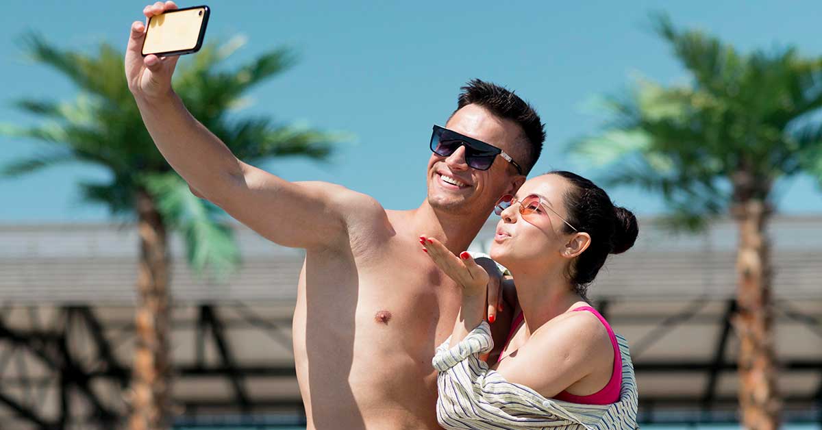Casal tirando selfie na praia, aproveitando dia ensolarado com palmeiras ao fundo, demonstrando felicidade e bom humor ao fazer fotos juntos.